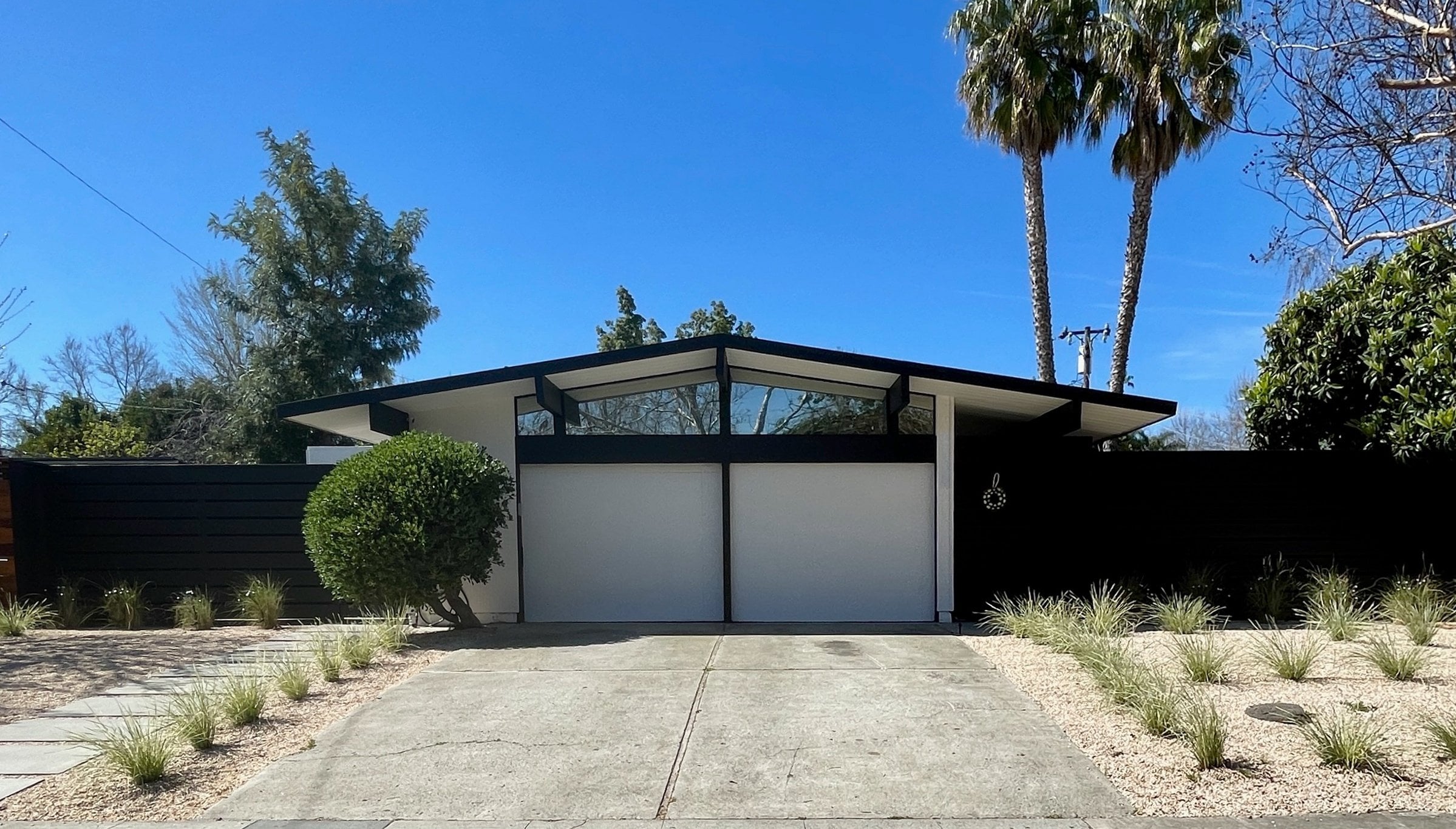 An Eichler home with a low gabled roof, clerestory windows, and a dark fence, under a clear blue sky